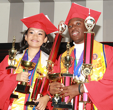 Two happy graduates in red cap and gown holding trophies